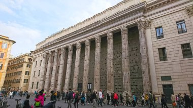 Roma, İtalya - 4 Nisan 2018: Tempio di Vibia Sabina e Adriano 'nun ayakta duran sütunlarının yanından geçen turistler, salgından önce bulutlu bir gökyüzüne karşı şehrin tarihsel özünü somutlaştırdılar.