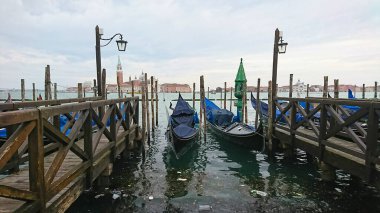 Venedik, İtalya - 9 Nisan 2018: Venedik 'in ikonik binaları arka planda belirirken, geleneksel gondolların denize yansıyan ahşap bir iskeleye demirlediği manzara