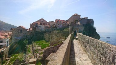 Dubrovnik, Hırvatistan - 14 Nisan 2018: Ortaçağ taş duvarları, açık mavi bir gökyüzü altında Adriyatik Denizi manzarasının keyfini çıkararak surlar boyunca yürüyen insanlarla tarihi kenti çevreledi