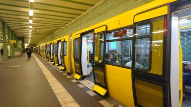 Berlin, Almanya - 18 Nisan 2018: Canlı sarı bir U-Bahn treni, modern mimari işlek kentsel geçiş ortamını çevreleyen yolcularla birlikte istasyonda duruyor.