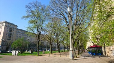 Berlin, Almanya - 4.18.2018: İlkbahar atmosferinde parlak mavi gökyüzü altında apartman binaları ile kaplı geniş bir yürüyüş yolu boyunca yemyeşil ağaçların yer aldığı sakin bir kent manzarası