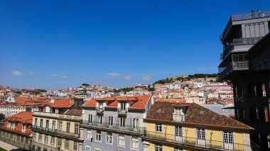 Lizbon, Portekiz - 2 Haziran 2018: Lizbon ve Castelo de S. Jorge 'nin tepedeki kırmızı kiremitli çatılarının manzaralı manzarası, açık mavi ve güneşli gökyüzünün altında Elevador de Santa Justa