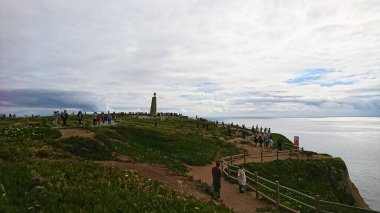 Cabo da Roca, Portekiz - 3 Haziran 2018: Atlantik Okyanusu 'nun yanındaki bulutlu gökyüzünün altındaki anıta hayran olan turistlerin yürüdüğü engebeli kıyı şeridi manzarası