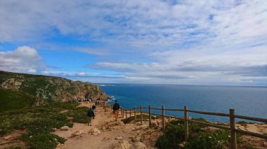 Cabo da Roca, Portekiz - 3 Haziran 2018: Avrupa 'nın en batı noktasına uzanan manzaralı kıyı yolu, ziyaretçiler canlı bir gökyüzü ve sakin bir denizin altında kayalık bir patika boyunca yürüyor
