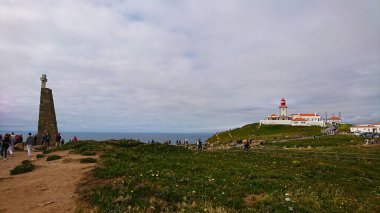 Cascais, Portekiz - 3 Haziran 2018: Cabo da Roca 'daki ikonik deniz feneri bulutlu bir günde Atlantik Okyanusu' na bakan turistler engebeli kıyı manzarası ve canlı kır çiçeklerinin keyfini çıkarıyorlar