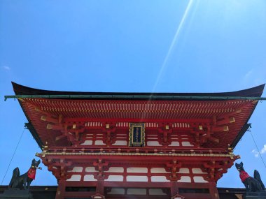Fushimi, Japonya - 08.08.2024: Fushimi Inari-taisha 'daki kule kapısı dekore edilmiş yapı ve süslenmiş ahşap bir levhanın üzerindeki tapınak ismiyle berrak mavi gökyüzü altında iki taş tilki heykelinin yanında.