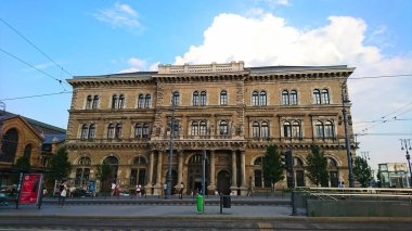 Budapeşte, Macaristan - 4 Haziran 2018: Açık mavi gökyüzünün altında sütunları olan Budapeşte Corvinus Egyetem binasının ön cephesinde, tramvay durağında bekleyen yayalar var.