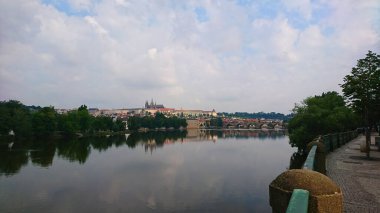 Prag, Çek Cumhuriyeti - 11 Haziran 2018: Vltava Nehri boyunca uzanan Prag Kalesi 'nin ikonik görünümü, tarihi mimari ve yemyeşil manzarayı bulutlu bir gökyüzü altında yansıtıyor