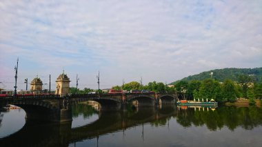 Prag, Çek Cumhuriyeti - 11 Haziran 2018: Majestic Vltava nehir köprüsü antik mimari, tramvaylar ve sabah çevresindeki yemyeşil alanlar ile sakin sulara yansıyor