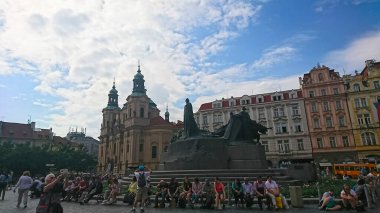 Prag, Çek Cumhuriyeti - 12 Haziran 2018: Turistler Eski Şehir Meydanı 'ndaki Jan Hus Anıtı' nın yanında dinlenirken, arkasında Aziz Nicholas Kilisesi, parçalı bulutlu gökyüzü altındaki tarihi binalarla çevrili