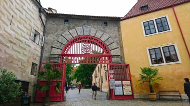 Cesky Krumlov, Çek Cumhuriyeti - 12 Haziran 2018: Cesky Krumlov Şatosu 'nun tarihi kırmızı tuğlalı kapısı sarı ve gri binalar çerçeveli, ziyaretçiler bulutlu bir gökyüzü altında kaldırım taşı patikasında geziniyorlar
