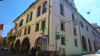 Münih, Almanya - 6.15.2018: İkonik Hofbrauhaus binası açık mavi gökyüzüne karşı canlı bir şekilde duruyor, ziyaretçiler Münih 'in kültür sahnesinin göbeğindeki canlı sokak atmosferinin keyfini çıkarıyorlar.