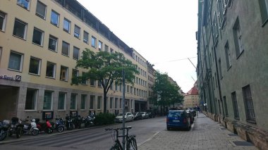 Münih, Almanya - 6.15.2018: Şehir merkezinde şehir yaşamını ve mimari çeşitliliği bulutlu gökyüzü altında gösteren modern binalar, ağaçlar ve park edilmiş araçlarla dolu sakin bir cadde