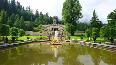 Bavyera, Almanya - 6.14.2018: Linderhof Sarayı 'ndaki büyük çeşme, ziyaretçilerin sakin park atmosferinin keyfini çıkarırken altın heykelleri ve canlı yeşillikleri yansıtıyor
