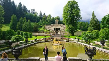 Bavyera, Almanya - 6.14.2018: Ziyaretçiler, yemyeşil ve dramatik dağ zirveleri tarafından çerçevelenen Schloss Linderhof bahçelerindeki altın çeşmenin yakınındaki anıları yakalıyorlar