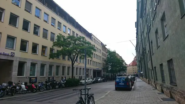 Münih, Almanya - 6.15.2018: Şehir merkezinde şehir yaşamını ve mimari çeşitliliği bulutlu gökyüzü altında gösteren modern binalar, ağaçlar ve park edilmiş araçlarla dolu sakin bir cadde