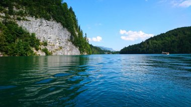 Konigssee, Almanya - 6.16.2018: Konigssee 'nin sarp kayalıklarla çevrili zümrüt sularının sakin manzarası ve berrak mavi gökyüzü altında süzülen bir tekne