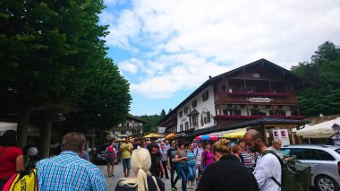 Konigssee, Almanya - 6.16.2018: Güneşli bir günde geleneksel Bavyera binaları ve yemyeşil ağaçlarla çevrili pitoresk bir pazar caddesinde gezinen turist manzarası