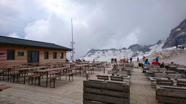 Zugspitze, Almanya - 6.17.2018: Geniş tepeler ve bulutlarla çevrili ahşap teraslı bir dağ kulübesi, ziyaretçileri ferahlatıcı bir dağlık deneyime davet ediyor