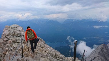 Zugspitze, Almanya - 6.17.2018: Parlak turuncu ceketli bir yürüyüşçü, bulutlu gökyüzünün ortasında nefes kesen zirveleri ve gölü göstererek kayalık arazide geziniyor