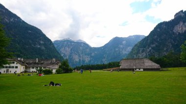 Berchtesgaden, Almanya - 6.16.2018: Ziyaretçiler, görkemli zirveler ve pitoresk bir dağ vadisindeki geleneksel dağlık binalarla çevrili yemyeşil bir çimenlikte dinleniyor