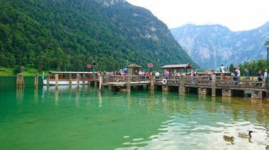 Konigssee, Almanya - 6.16.2018: Turistler, bulutlu bir yaz gününde turkuaz gölüne yansıyan yeşil dağlarla, alp sularına tepeden bakan hareketli bir ahşap iskelede toplandılar