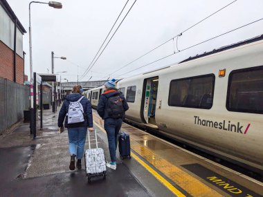 St Albans, İngiltere - 1.2025: Yuvarlanan bavullu iki yolcu, bulutlu bir gökyüzü altında modern bir Thameslink treninin yanında ıslak bir platform boyunca yürüyor.