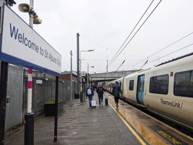 St Albans, İngiltere - 1.2025: Bavul taşıyan yolcular, bulutlu bir gökyüzü altında, St Albans City tren istasyonundaki sabit Thameslink treninin yanında, 4 numaralı ıslak platform boyunca yürürler.