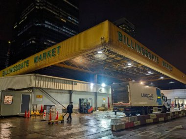 Londra, İngiltere - 1.25.2025: Bir işçi, gece Billingsgate Market 'in aydınlık giriş tepe örtüsünün altında, ıslak kaldırımı yansıtan bir yük kamyonunun yanında palet krikosu çekiyor.