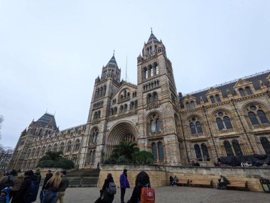 Londra, İngiltere - 2.15.2025: Doğa Tarihi Müzesi 'nin bulutlu bir gökyüzü altında dekore edilmiş ön cephesi, ziyaretçiler giriş alanının etrafında dolanıp adımlar atıyorlar