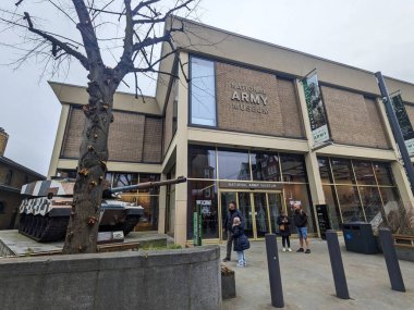 Londra, İngiltere - 2.15.2025: Ziyaretçiler Chelsea 'deki Ulusal Ordu Müzesi' nin modern cam girişinde toplanarak, bulutlu bir gökyüzü altında bir askeri tank sergisi ve çıplak kış ağacı yer alıyor.
