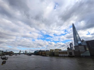 Londra, İngiltere - 3.16.2025: Thames Nehri üzerinde dramatik bulutlu gökyüzü, The Shard skyscraper, Tower Bridge ve nehir trafiğiyle ikonik şehir silueti sergileniyor