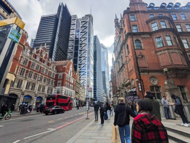 Londra, İngiltere - 316 2025: İnsanlar işlek bir caddenin yanındaki kaldırımda yürüyor modern gökdelenlerle kırmızı tuğlalı binalar ve bulutlu bir gökyüzü altında kırmızı çift katlı bir otobüs arasında.