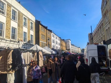 Londra, İngiltere - 3.29.2025: Alışverişçiler, Notting Hill 'deki ünlü Portobello Road Market' inde güneşli bir günde geleneksel Londra mimarisine karşı açılan renkli giyim tezgahlarına bakıyorlar