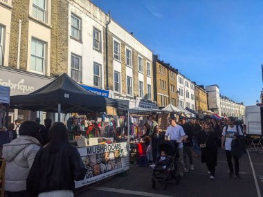Londra, İngiltere - 3.29.2025: Canlı kalabalık, güneşli bir günde geleneksel Londra mimarisine karşı kalabalık bir açık hava sokak pazarında yiyecek ve hediyelik eşya satan tezgahlara göz atıyor