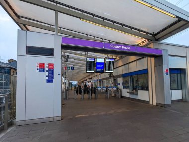 Londra, İngiltere - 2 Ağustos 2024: Yolcular, Custom House istasyonunun modern girişindeki bilet engellerini, Elizabeth Line ve DLR kavşak merkezlerini ve programlarını gösteren dijital ekranları kullanıyorlar