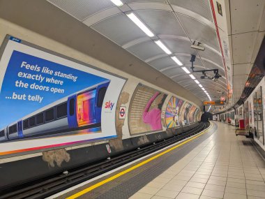 Londra, İngiltere - 9 Kasım 2024: Dokunsal asfalt ve reklamlarla dolu boş bir kavisli platform, Shepherds Bush Londra metro istasyonunda bir tünele giden yollar.