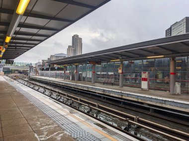 Londra, İngiltere - Aralık 12024: Stratfords modern DLR istasyonunda boş, ıslak bir platform ve raylar boyunca yapay ışıklar altında ve arka planda şehir mimarisi ile kapalı bir gökyüzü