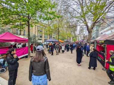 Londra, İngiltere - 4.27.2023: Kalabalık, Londra parkında bulutlu bir gökyüzünün altındaki yapraklı bahar ağaçlarının altında renkli tezgahları olan canlı bir sokak gıda pazarında toplandı