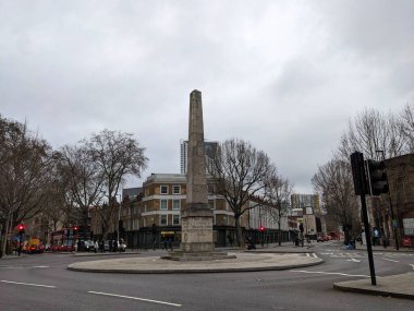 Londra, İngiltere - 28 Şubat 2023: Tarihi St Georges Circus obelisk, bulutlu bir kış gökyüzü altında, kırmızı bir otobüs de dahil olmak üzere yoğun bir kavşağın merkezinde yer alıyor.