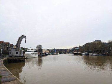 Bristol, İngiltere - 29 Ocak 2025: Bir kentsel limandaki modern bina ve teknelerin yanında tarihi bir liman vinci, sanayi mirası ve şehir hayatının bir karışımını bulutlu bir gökyüzü altında yansıtıyor