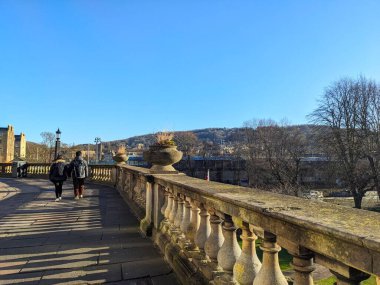 Bath, İngiltere - 30 Ocak 2025: Güneşli bir günde Avon Nehri 'ne ve şehrin klasik mimarisine tepeden bakan tarihi bir taşın üzerinde yürüyen iki kişi
