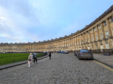 Bath, İngiltere - 30 Ocak 2025: Turistler, yeşil çimlere ve kaldırım taşı sokağına bakan bulutların altında, Gürcistan Kraliyet Hilal 'inin sembolik ön cephesini ziyaret ederek fotoğraflarını çekiyorlar.