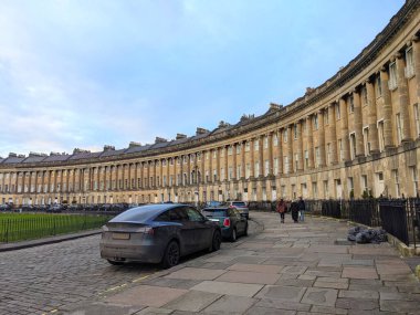 Bath, İngiltere - 30 Ocak 2025: Yayalar geniş kaldırım ve kaldırım taşı caddeleri boyunca Kraliyet Hilal 'inin ikonik eğimli Gürcistan konakları önünde bulutlu bir gökyüzü altında yürüyor