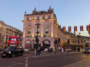 Londra, İngiltere - 20 Haziran 2022: Kırmızı çift katlı bir otobüs ve siyah bir taksi Piccadilly Sirki 'nin işlek caddesinde aydınlık binalar ve dalgalı bayraklar altında geziniyor