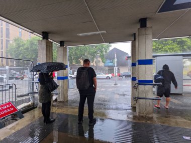 Londra, İngiltere - 15 Ağustos 2022: Ani sağanak yağış sırasında insanlar Blackhorse Road Station 'da beton bir çatı altında sığınak arıyor, şehir caddesini bulanıklaştırıyor ve arabalar geçerken su birikintileri yaratıyor