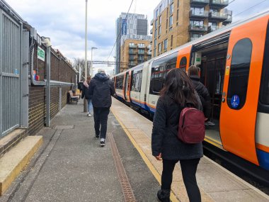 Londra, İngiltere - Nisan 01 2022: Çeşitli yolcular bulutlu bir gökyüzü altında Walthamstow Merkez tren istasyonunda turuncu bir Londra metro istasyonunun yanından geçtiler