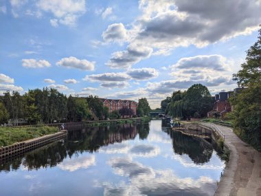 Londra, İngiltere - 19 Eylül 2021: Yapılar ve ağaçlar, bulutlu gökyüzünü yansıtan huzurlu Lea Nehri 'nin hizasında, insanlar sahil boyunca yürüyorlar.