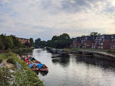 Londra, İngiltere - 18 Eylül 2021: Bulutlu bir gökyüzünün altındaki konut binaları ve ağaçlarla kaplı sakin Lea Nehri boyunca renkli dar kayıklar demirlemiştir