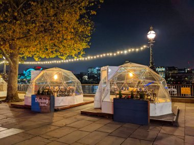Londra, İngiltere - 21 Aralık 2021: Gece Thames Nehri boyunca güney yakasında büyük bir ağaç ve sokak lambası yanında açık havada yemek için aydınlatılmış iç mekanları olan saydam jeodezik yemek kubbeleri